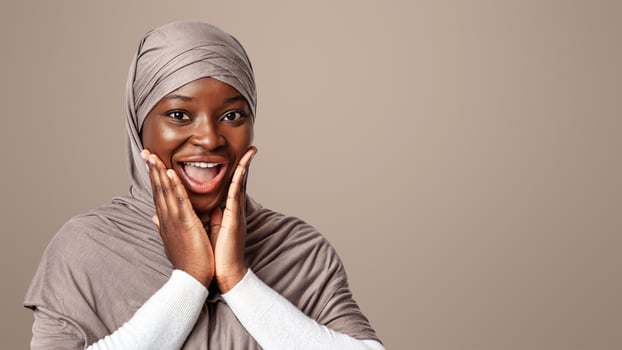 Surprised Black Woman In Hijab Touching Face In Excitement, Joyful African Muslim Lady Emotionally Reacting To Good News, Standing Over Light Studio Background, Looking At Camera, Copy Space photo