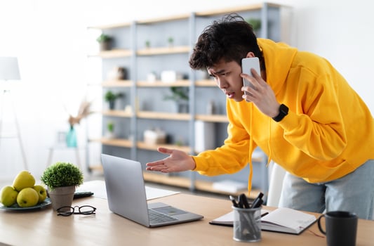 Confused Discontented Asian Man Talking On Phone Using Laptop Having Problem With Computer Or Internet Connection At Home. Displeased Male Customer Calling To Hotline Service And Complaining photo