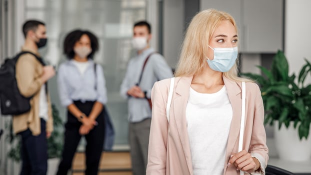 Workers in modern office hallway return to work after quarantine. Focus on beautiful blonde woman in business suit and protective mask looks to the side and blurred background, empty space photo