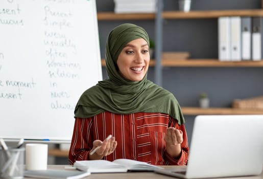 E-learning Concept. Smiling female arabic teacher in headscarf making call, explaining English grammar rules to students. Young muslim woman talking and looking at pc screen, sitting at desk photo