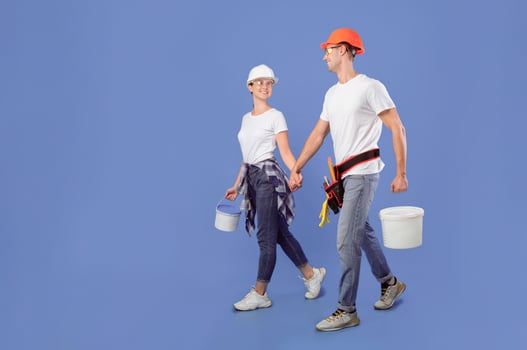 Young spouses in safety glasses and hardhats walking with paint equipment over blue background, millennial workers couple ready for apartment repair and house renovation, full length shot, copy space photo