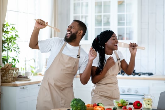 Kitchen Concert. Cheerful Black Couple Having Fun While Cooking Together At Home, Positive African American Spouses Wearing Aprons Singing And Dancing, Using Spatulas As Microphones, Free Space photo