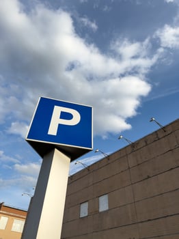 Tall urban parking sign with bold white letter P on blue square background, mounted on pole against cloudy blue sky, with beige brick building and architectural lighting fixtures in background photo