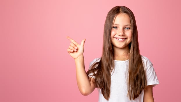 Kid with long hair presenting product. Happy little girl shows to empty space, isolated on pink background, studio shot photo