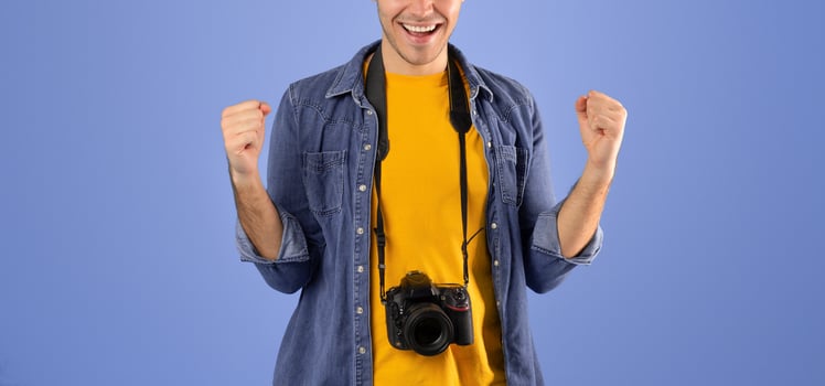 Cheerful male tourist with professional camera making YES gesture over blue studio background. Talented amateur photographer with modern equipment ready to take pictures during his travels photo