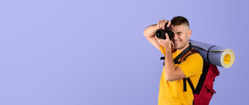 Cheerful young traveler with backpack and camera ready to take photos on blue background
