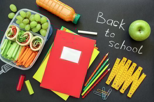 Arrangement of healthy snacks including wraps, grapes, and carrots alongside school supplies like colored pencils and notebooks on black chalkboard, signaling back to school season. photo