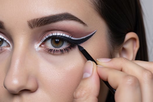 Extreme close-up of a makeup artist skillfully applying bold white and black graphic winged eyeliner to a woman's eye. High-fashion, avant-garde beauty, and cosmetic artistry concept photo
