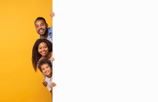 Cheerful black family of three peeking out from behind white advertisement board, happy parents and preteen son standing against yellow background, recommending new offer, copy space photo