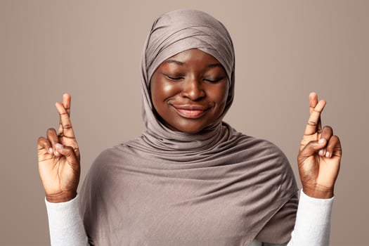 Superstitious Black Muslim Woman In Hijab Making Wish With Crossed Fingers And Closed Eyes, Smiling African Islamic Lady Pleading For Luck And Fortune Over Light Studio Background, Copy Space photo