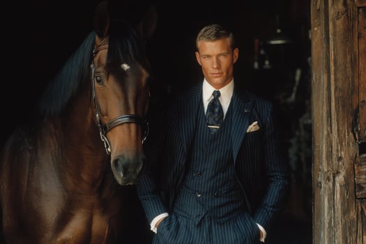 A man in a tailored pinstripe suit stands confidently beside a brown horse inside a rustic barn. photo