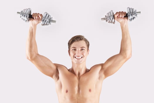 Portrait of positive young athlete lifting dumbbells above his head and smiling on light studio background. Strong bodybuilder with six pack abs working out his biceps, pumping iron photo