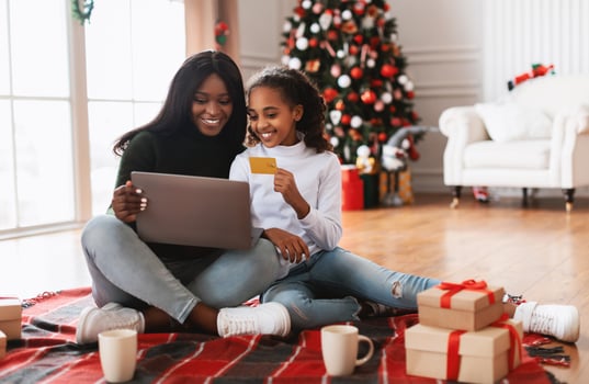 Christmas Preparation. Smiling black girl and lady using pc notebook, choosing, ordering and buying gifts online, cheerful teen daughter holding golden credit card in decorated living room interior photo