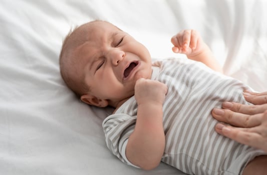 Closeup Portrait Of Crying Little Newborn Baby In Bodysuit Lying On Bed At Home, Upset Infant Child Suffering Colics Or Gas Problems, Loving Mother Making Tummy Massage To Small Kid With Two Hands photo