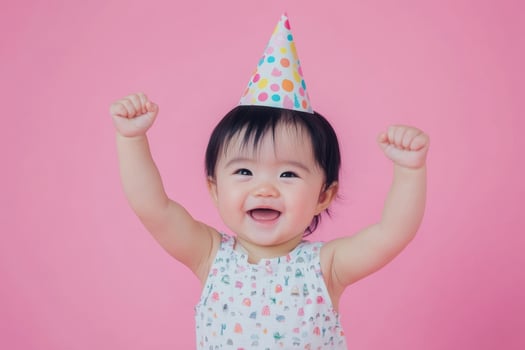 Baby with black hair celebrating in a romper and party hat against a pink background photo