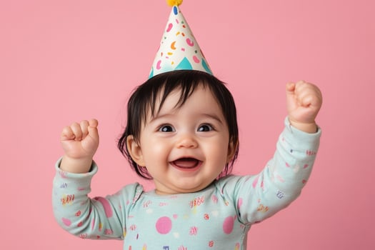 Excited baby with black hair in a romper celebrating with a party hat against a pink background photo