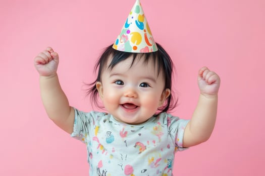 Happy baby with black hair wearing a party hat and celebrating against a pink background photo