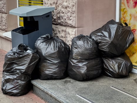 Black garbage bags are stacked next to a public trash bin outside a shop on a busy city sidewalk during the afternoon photo