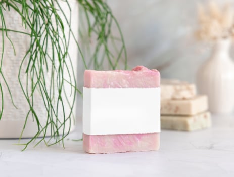 Soap bar with blank label on marble near green plant close up, cosmetic mockup photo