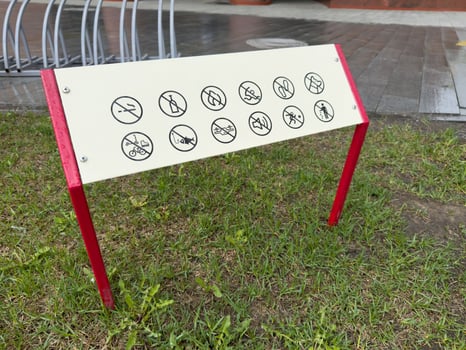 Prohibition sign with multiple black icons on a white board supported by red legs, installed on a lawn next to a walkway, indicating banned activities in a public space for safety and order photo