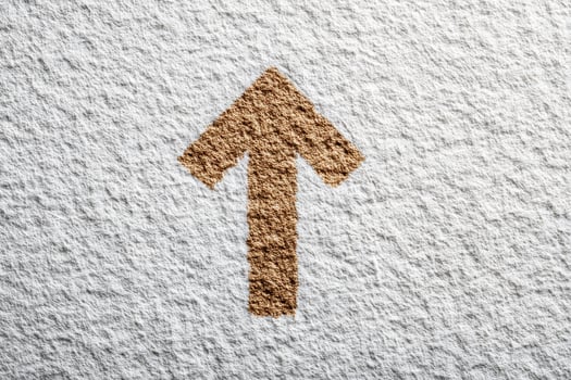 A tan arrow points upward. The arrow and background surface have a rough, textured appearance. The arrows color contrasts with the white, uneven surface. photo