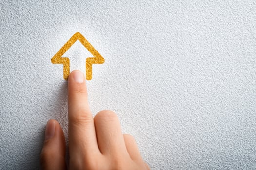 A hand is positioned to touch a stylized golden arrow emblem on a light textured wall. The focus captures the interaction between the human element and the graphic design. photo