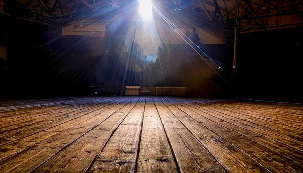 A worn wooden stage stretches into darkness, a single spotlight blazing at center stage. Long shadows spill across the floorboards photo