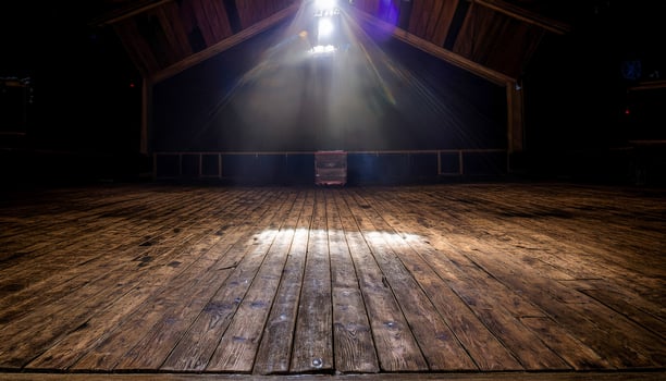 Empty wooden stage floor illuminated by a bright spotlight from above, with visible rafters and a dark backdrop. Evokes anticipation and the potential for performance in a rustic setting. photo