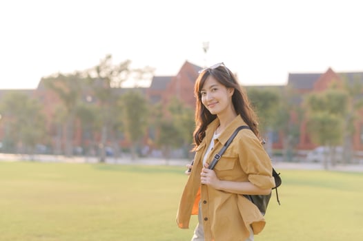 Southeast Asian woman enjoying travel outdoors with cheerful smile, wearing casual clothing and backpack, standing on green grass with sunlight in background photo