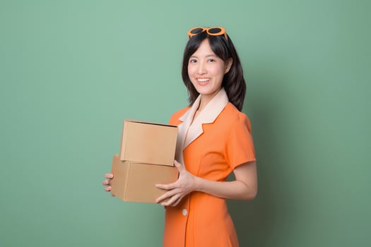 Southeast asian woman in orange dress holding cardboard boxes with cheerful expression standing against green background, representing delivery or online shopping concept photo