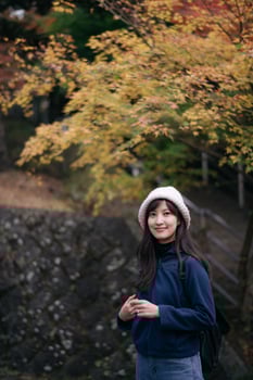 Asian woman in blue sweater and white hat smiles by autumn foliage, enjoying outdoor holiday in Kyoto, cheerful and relaxed mood photo