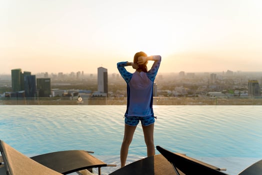 Woman standing by rooftop infinity pool at sunset, tying her hair while enjoying panoramic city skyline view and golden hour light in peaceful atmosphere photo
