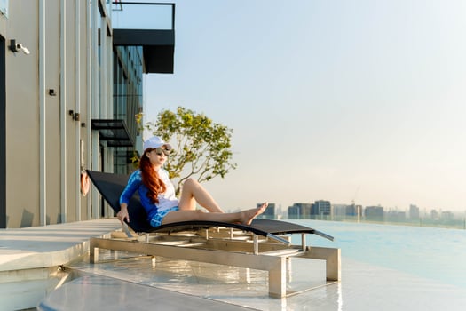 Young woman in sunglasses and sporty swimwear relaxing on a lounge chair by rooftop infinity pool, enjoying peaceful sunny day with panoramic city view photo