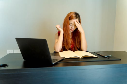 Stressed young Asian woman feeling pressure while studying or working from home, holding her head in frustration at desk with laptop and notebook. photo