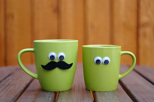 Two green coffee mugs decorated with googly eyes and mustache on a wooden table background photo