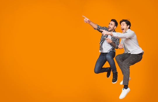 Overjoyed guys pointing at free space and jumping over orange studio background photo