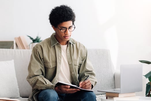 Studying from home concept. Black teen guy with laptop communicating online with teacher, taking notes during lecture. Funky university student writing down information on webinar photo