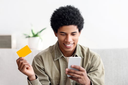 Cool African American teenager with laptop using mobile phone and credit card for online shopping at home. Funky black teen guy purchasing goods on web with help of cellphone photo