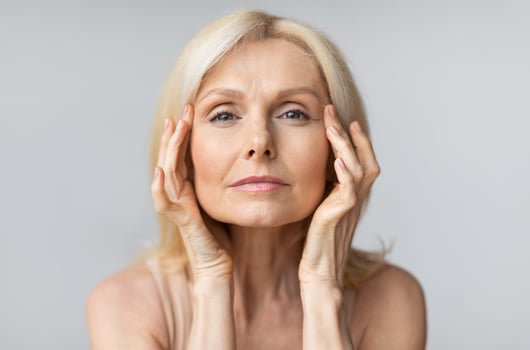 Antiage skincare. Senior woman touching skin near eyes and looking at camera, lady with flawless skin standing over grey studio background, closeup portrait photo