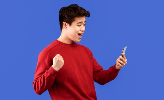 Yes, Big Luck. Portrait Of Excited Asian Guy Celebrating Triumph, Using Mobile Phone, Emotional Young Man Having Success, Holding Cellphone, Raising And Shaking Clenched Fist, Blue Studio Background photo
