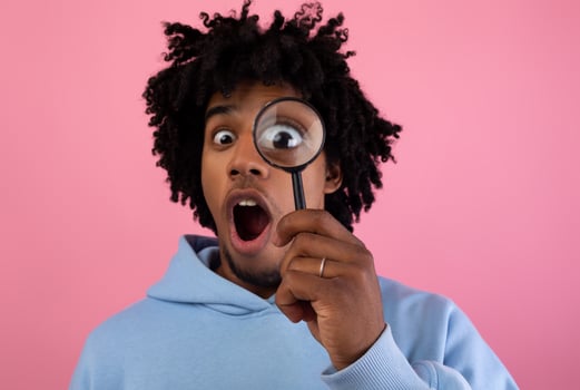 Shocked black teen guy looking through magnifying glass on pink studio background. Surprised African American teenager suspecting, investigating or questioning something. Secret, doubt concept photo