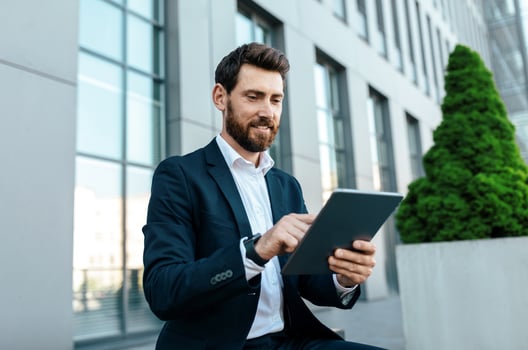 Smiling young handsome european businessman with beard in suit typing on tablet near modern office building, outdoor. Chat in new app, manager work with technology and new normal, business in city photo