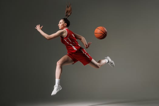 Dynamic female basketball player in mid-air jump with flying ball, wearing red uniform on gray studio background symbolizing strength, motion, and athletic performance. photo