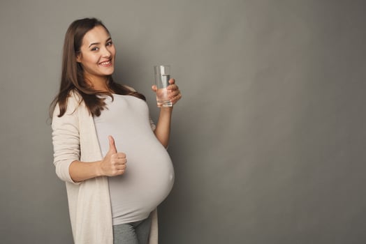Pregnant woman with water showing thumb up gesture. Young expectant lady have rest with glass, gray studio background, copy space. Pregnancy, healthcare, thirst concept photo
