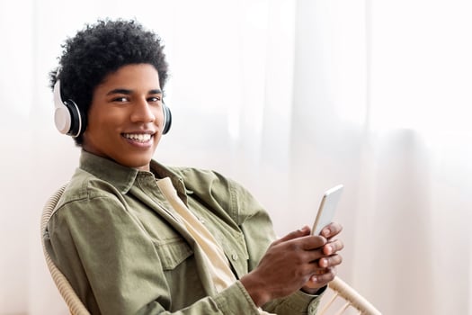Handsome black adolescent in headset picking music playlist on mobile phone at home. Cool African American teen guy listening to his favorite song, audiobook or radio on cellphone photo
