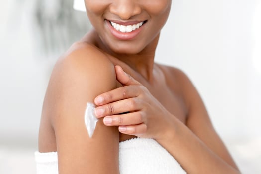 Organic skin care, routine and moisturizing products. Smiling young african american lady blogger in white towel after shower applies cream on hand, in interior of bedroom in morning, copy space photo