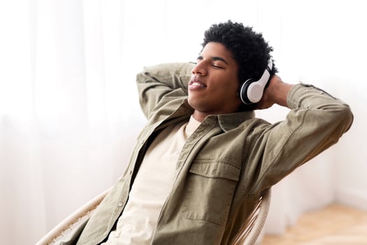 Cheerful black teen guy in headset relaxing to his favorite music while sitting in comfy armchair, panorama. Peaceful African American youth listening to online radio channel at home photo
