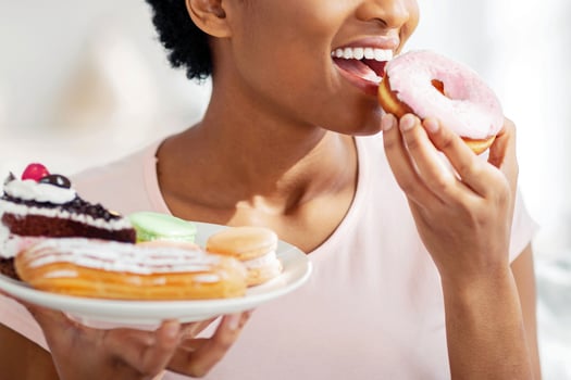 Diet breakdown, cheat meal and imbalanced nutrition concept. Beautiful young black woman with plate of sweets eating unhealthy donut at home. African American lady eating junk food photo