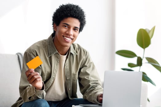 Cybershopping concept. Smiling black teenage guy with laptop and credit card ordering goods online from home. Happy African American teen purchasing things on web indoors photo