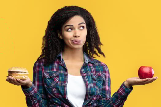Healthy Vs Unhealthy Food Concept. Puzzled Black Lady Holding Burger And Apple Fruit. Hungry African American Woman Choosing What To Eat, Posing Isolated Over Orange Studio Background. Banner photo